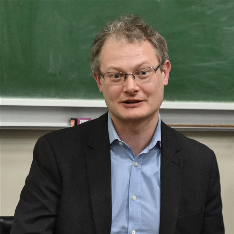 A photo of Dr Andrew Dilley with a blue shirt, black jacket and glasses against a green blackboard background