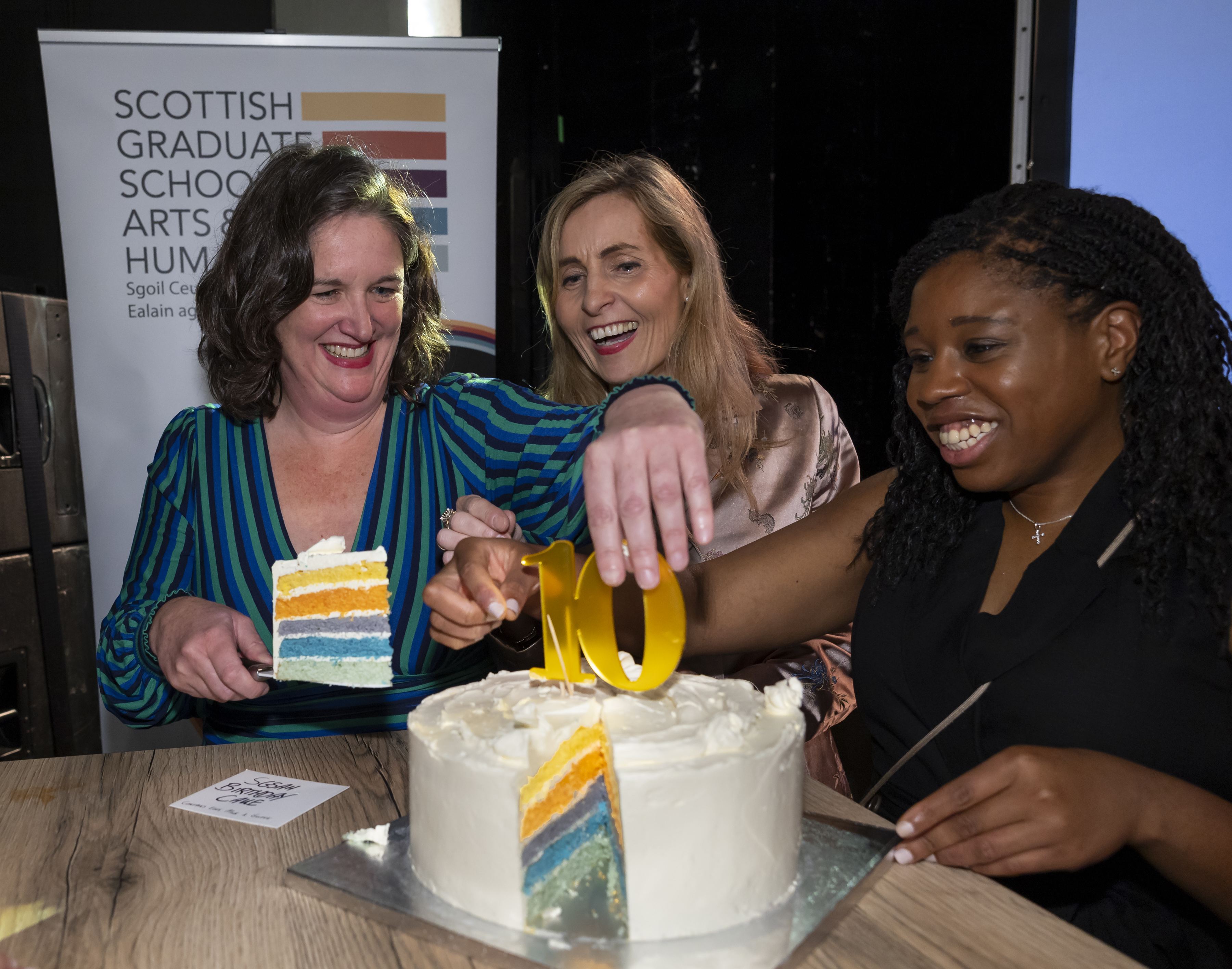 3 people smiling and cutting a rainbow cake