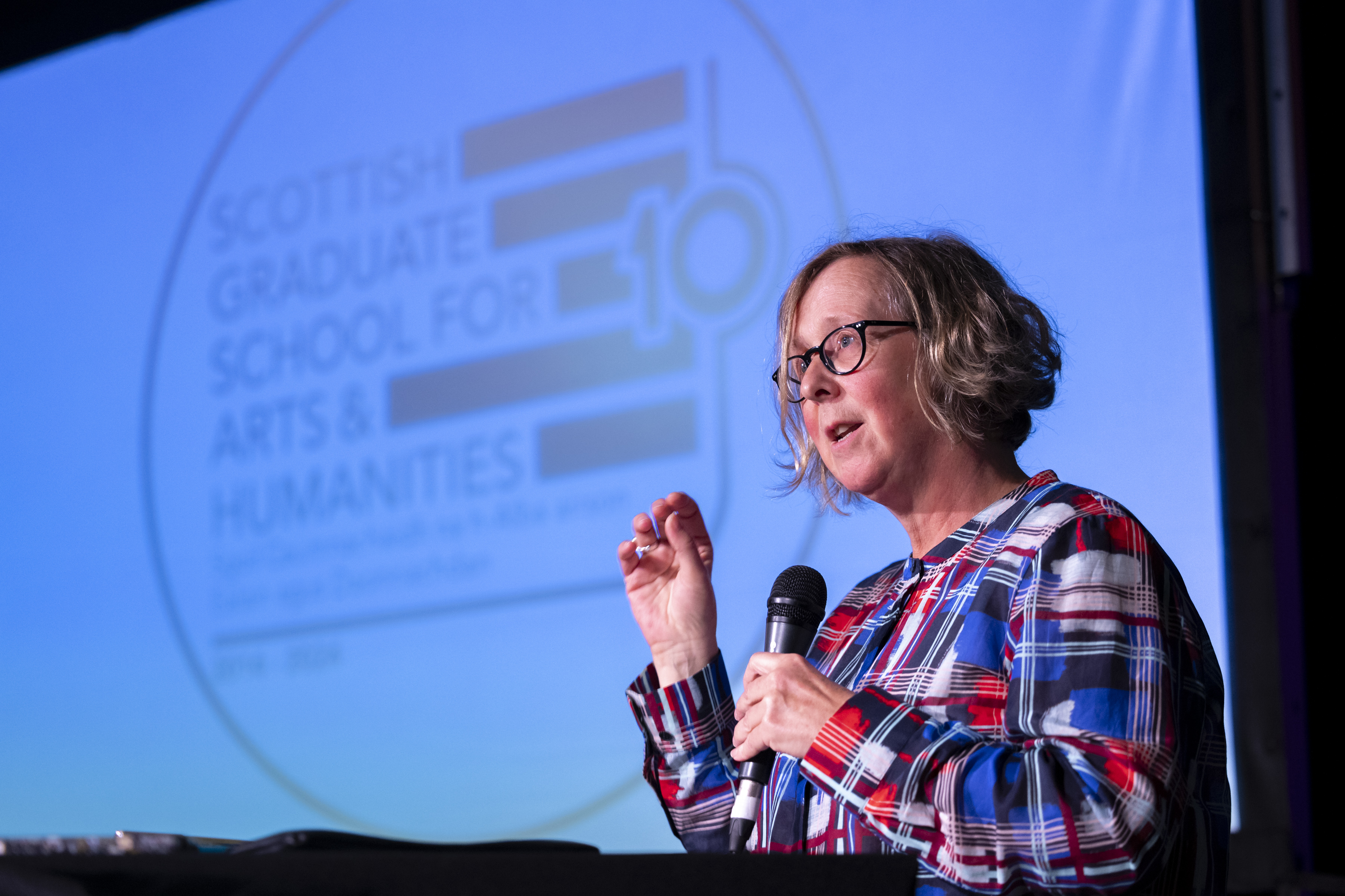 Professor Dee Heddon holding a microphone in front of a gold SGSAH logo on a screen