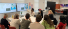 Participants seated around a table in a workshop setting, listening to a presenter who points to slides displayed on two large screens
