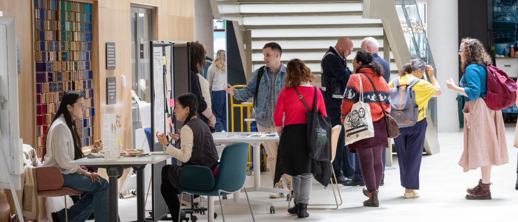 People browse and discuss research displays in a bright atrium, with poster boards and exhibition tables set up beneath a large staircase.