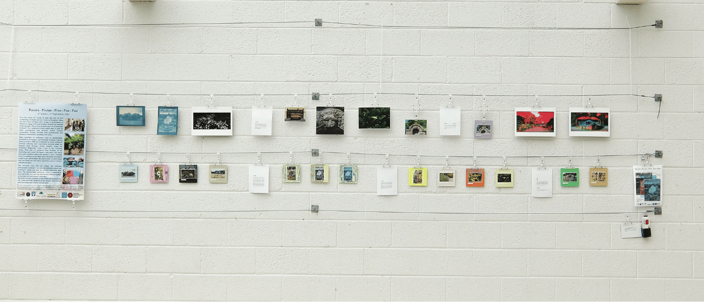 Exhibition display of small artworks and photographs arranged in rows on a white gallery wall