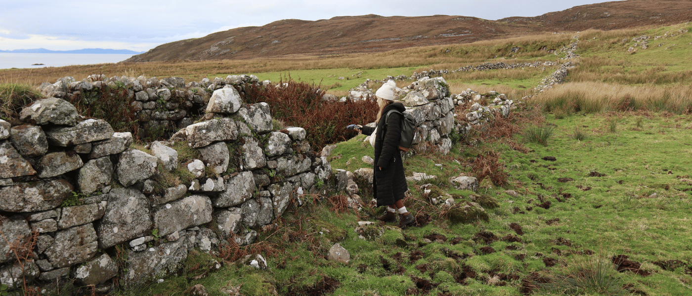 A researcher standing beside the remains of a stone structure in a grassy rural landscape