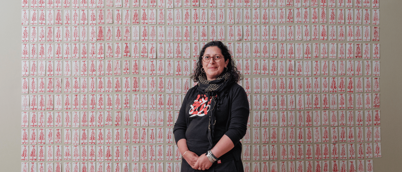 An artist-researcher standing in front of a large wall installation composed of hundreds of small white cards printed with red botanical motifs, arranged in a grid pattern