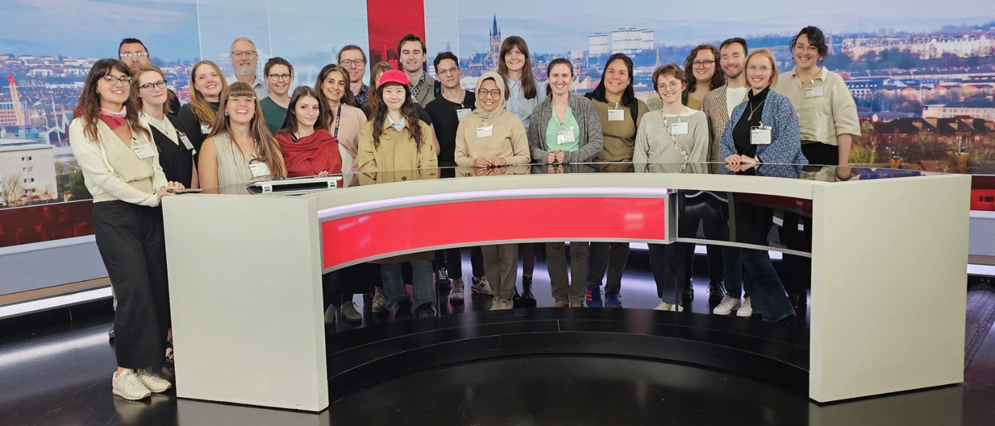 Group of workshop attendees standing behind a BBC Scotland news desk during a training session in a television studio, with a cityscape backdrop visible behind them.