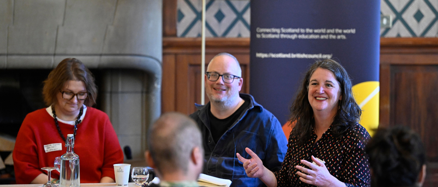 Participants seated around a table during a discussion at a workshop, with speakers addressing the group and a branded banner in the background