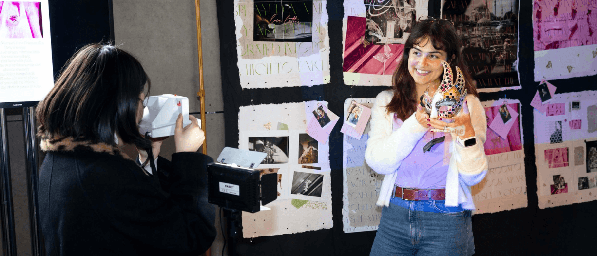 Person holding an ornate mask while being photographed in front of a display of artwork and photographs